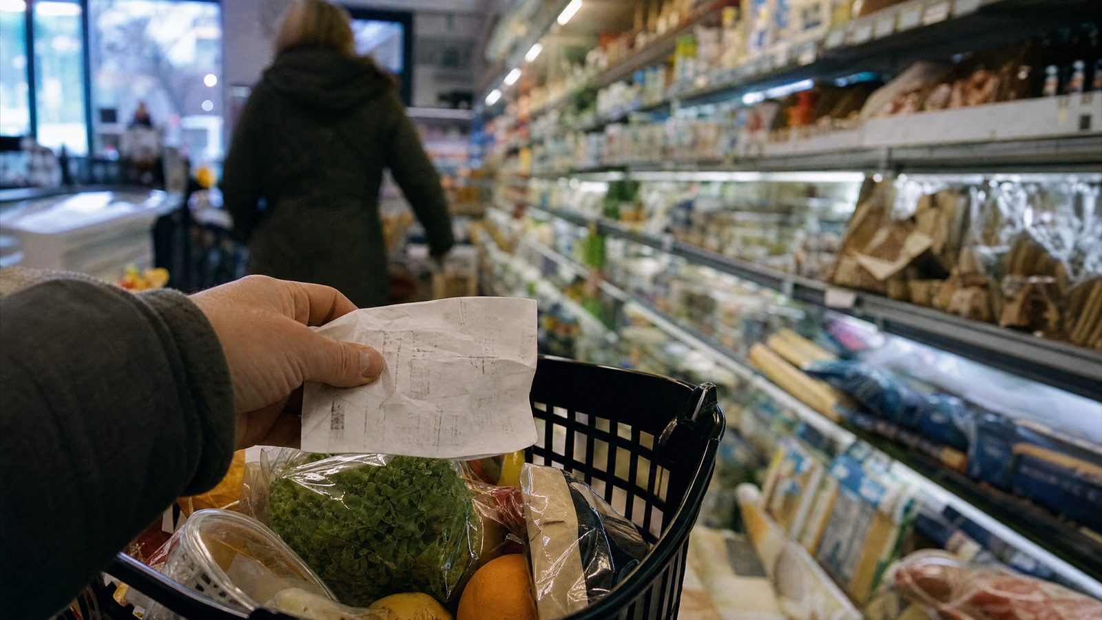 Mão a segurar um recibo de papel amarrotado sobre um cesto de compras num corredor de supermercado ao final da tarde.