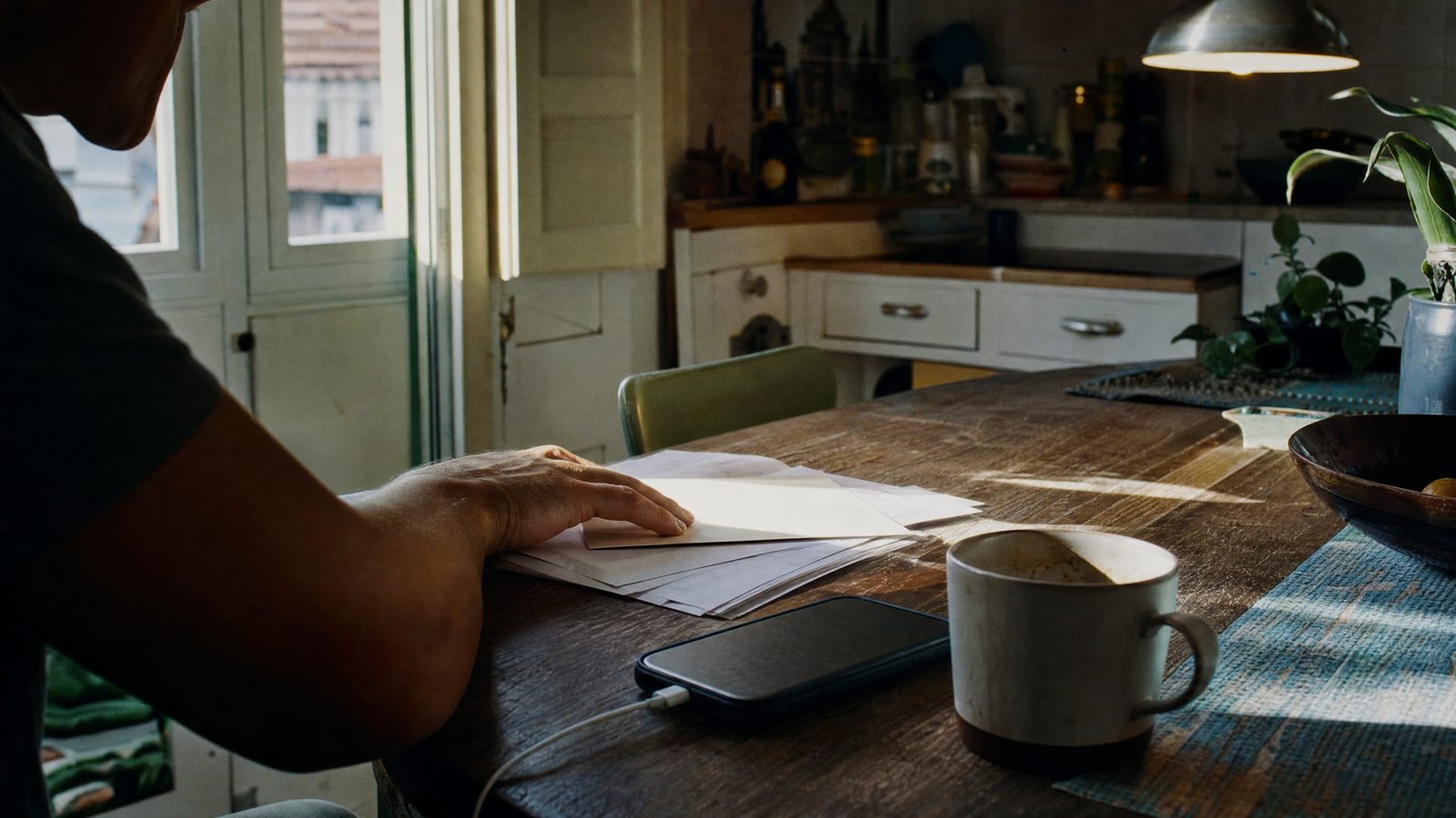 Uma pessoa em casa revê extratos e papéis financeiros numa mesa de cozinha, com luz da manhã a entrar pela janela.