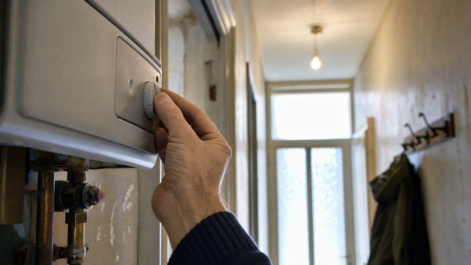 Mão a ajustar o termóstato de uma caldeira a gás num corredor de um prédio antigo